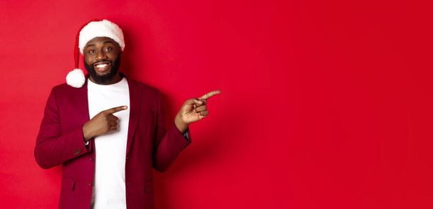Рождество, вечеринки и праздники. Улыбающийся чёрный мужчина с бородой и шляпой Санты, указывающий пальцами на логотип, стоящий на красном фоне. - Фото, изображение