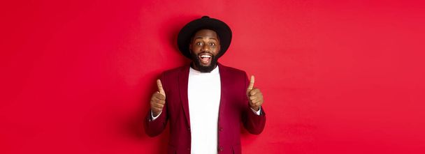 Christmas shopping and people concept. Handsome Black man smiling satisfied, showing thumbs-up, like and agree, approve something, standing against red background. - 写真・画像