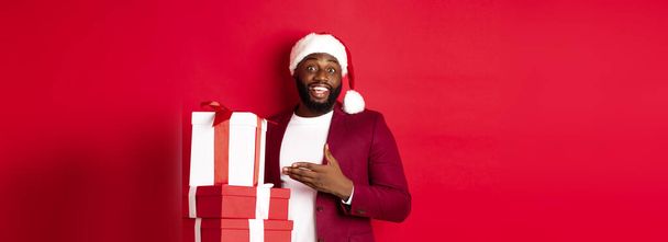 Χριστούγεννα, Πρωτοχρονιά και ιδέες για ψώνια. Χαρούμενος Μαύρος άντρας μυστικό Σάντα κρατώντας δώρα Χριστούγεννα και χαμογελώντας ενθουσιασμένος, φέρει δώρα, στέκεται ενάντια στο κόκκινο φόντο. - Φωτογραφία, εικόνα