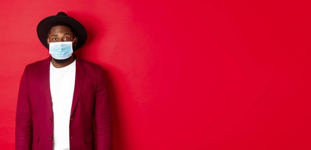 Covid-19 and fashion concept. Handsome african american man in stylish blazer and hat, wearing medical mask from coronavirus, standing over red background. - Foto, immagini