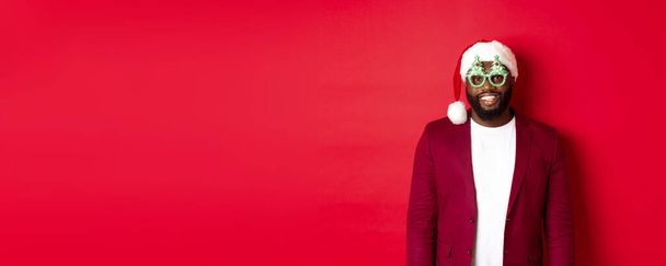 Grappige zwarte man in santa hoed en feestbril vieren Kerstmis, glimlachen gelukkig en wensen vrolijk Kerstmis, staan over rode achtergrond. - Foto, afbeelding