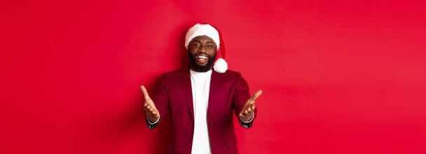 Χριστούγεννα, πάρτι και γιορτές. Ευτυχισμένος Μαύρος άνδρας με καπέλο Σάντα σας καλωσορίζω, δείχνοντας τα χέρια στην κάμερα με χαρούμενο χαμόγελο, στέκεται πάνω σε κόκκινο φόντο. - Φωτογραφία, εικόνα