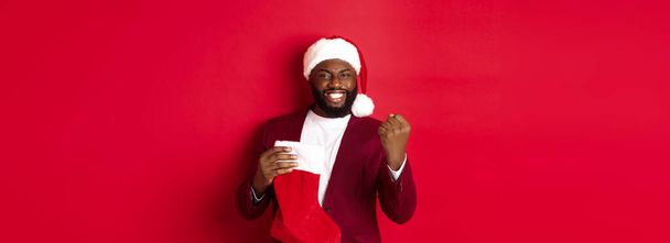Glücklicher Mann, der sich freut, Geschenke in Weihnachtssocke entgegennimmt, die Faust pumpt und zufrieden lächelt, vor rotem Hintergrund steht. - Foto, Bild
