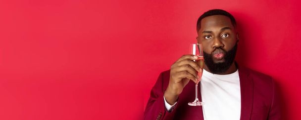 Közelkép vicces fekete férfi kóstoló pezsgő üvegből, néz buta és ráncos ajkak, ünneplő újév, álló ellen piros háttér. - Fotó, kép