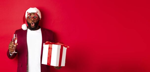 Karácsonykor. Jóképű afro-amerikai férfi party szemüveg és Mikulás kalap, kezében újévi ajándék és üveg pezsgő, kívánva boldog ünnepeket, piros háttér. - Fotó, kép