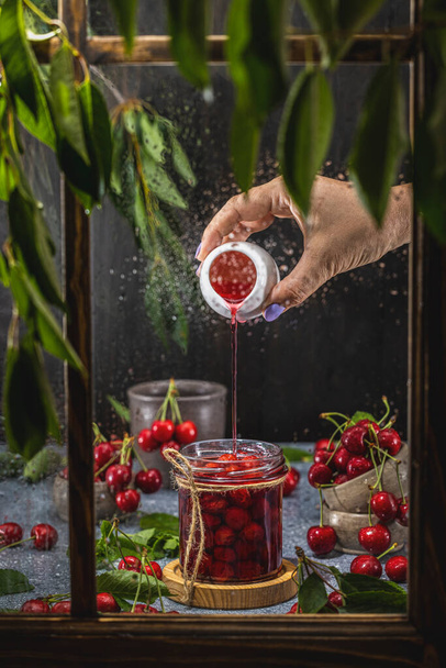 Caseiro DIY natural enlatado vermelho cereja geleia confiture com pimenta em frasco de vidro em pé sobre mesa de madeira escura atrás da janela - Foto, Imagem