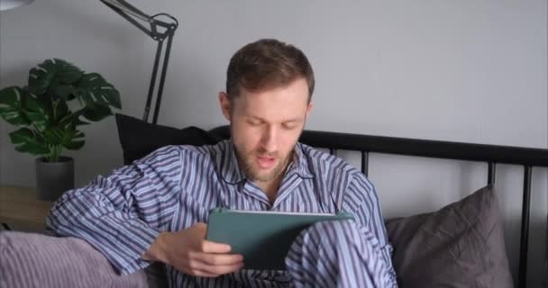Tevreden, eens emotie of Ja concept. Jonge bebaarde blanke man die 's ochtends nachtkleding draagt met een tablet in bed om te winkelen, te werken of nieuws te lezen. 4k-beelden - Video