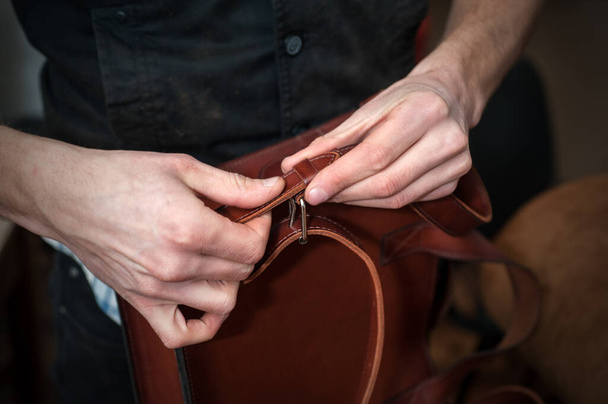 a craftsman in a workshop attaches a shoulder belt to a ready-made handmade leather briefcase - Foto, imagen