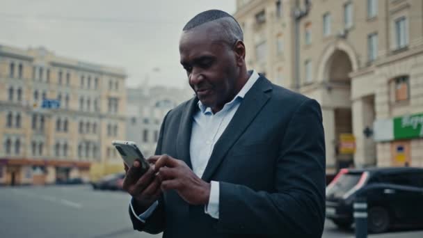 Афроамериканець, дорослий бізнесмен середнього віку, який працює в місті, дивлячись на телефон, читає хороші новини, отримує мобільне повідомлення, що пропонує онлайн-досягнення посміхаючись з успіхом смартфона. - Кадри, відео
