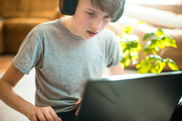 Diş teli takan bir genç dizüstü bilgisayarda yerde oturuyor. Kış bahçesinden gelen yeşil bitkilerin yanında şık iç mekanlarda çalışan çocuk evde online ders veriyor. - Fotoğraf, Görsel