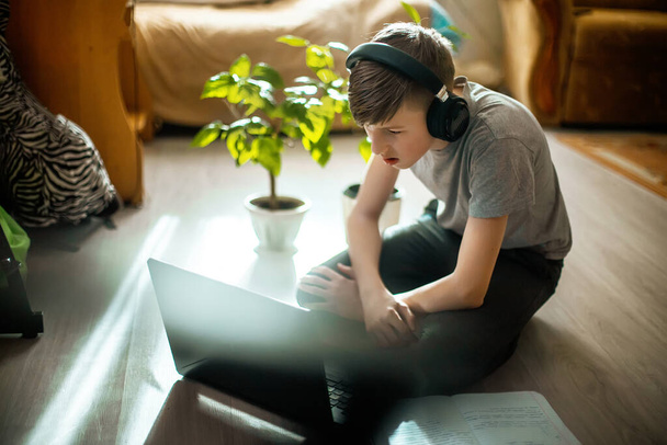 teenager in braces on teeth programs sitting at laptop on floor. boy in fashionable interior near trendy green plants from winter garden teaches lessons online at home - Foto, Imagen