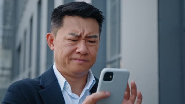 Close up angry worried stressed Asian middle-aged 40s man user businessman employer entrepreneur trying browsing on broken cellphone outdoors in city annoyed with mobile app error connection problem - Footage, Video