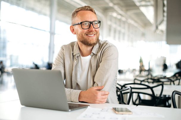 Positiv intelligenter erfolgreicher kaukasischer Mann mit Brille, in stylischer Freizeitkleidung, IT-Spezialist, Firmenseo, Freiberufler, sitzt am Schreibtisch mit Laptop beim Coworking, schaut weg, lächelt glücklich - Foto, Bild