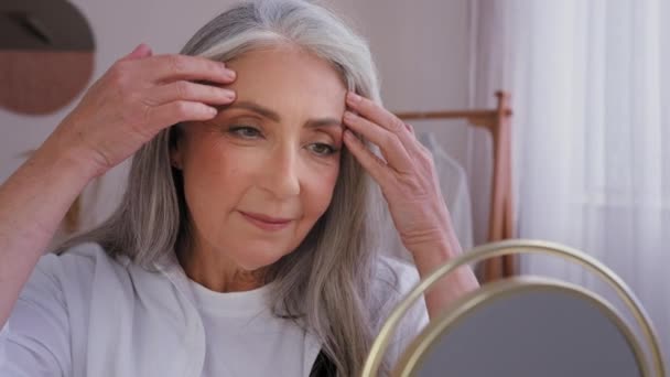 Крупный план Кавказская пожилая женщина 50-х годов женщина смотрит на зеркальное отражение массирующий лоб йога лицо упражнения гладкие морщины анти-морщинистый массаж касаются анти-старения поднимая кожу уход - Кадры, видео