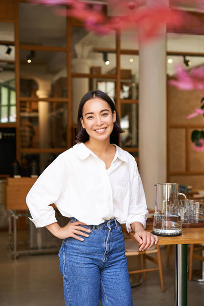 Glimlachende Aziatische manager, zelfverzekerde vrouw in de buurt van restaurant ingang, cafe eigenaar verwelkomt gasten. - Foto, afbeelding