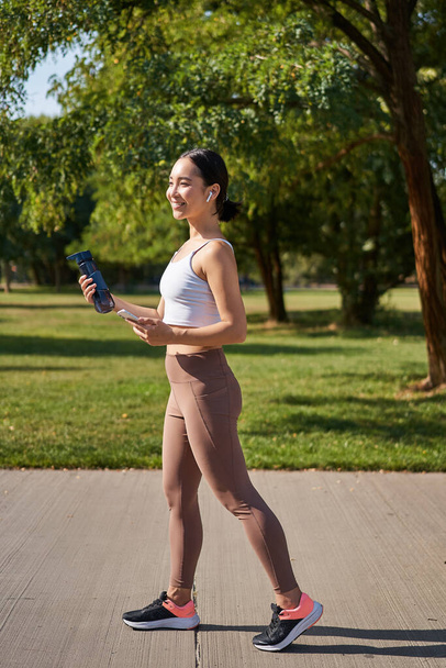 Actief Aziatisch meisje, in fitnesskleding, workout in het park, wandelen in sportkleding met smartphone en waterfles. - Foto, afbeelding
