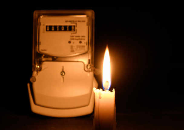 Κερί στο χέρι λάμπει στο σκοτάδι κοντά στο μετρητή ηλεκτρικής ενέργειας κατά τη διάρκεια διακοπής ρεύματος στο σπίτι. Πόλη, χώρα χωρίς ηλεκτρικό ρεύμα - Φωτογραφία, εικόνα