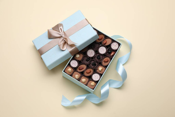 Open box of delicious chocolate candies and light blue ribbon on beige background, flat lay - 写真・画像