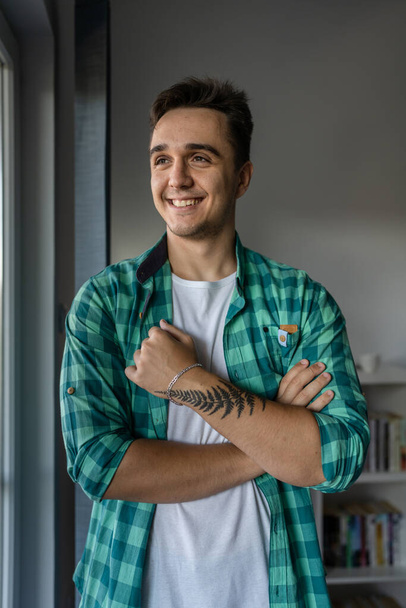 One man young adult male caucasian with brown hair standing by the window at home in day confident looking to the side happy smile wear green shirt real people copy space - 写真・画像