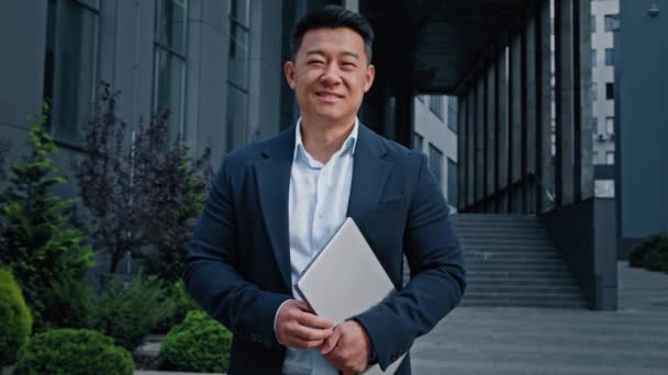 Asian 40s middle-aged businessman entrepreneur employer man worker manager designer financial analyst standing in city near office company building in downtown posing holding laptop looking at camera - Footage, Video