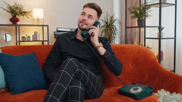 Портрет мужчины, ведущего телефонный разговор с друзьями, сидящими на диване в домашней комнате. Счастливый взволнованный молодой парень наслаждается старомодным ретро-телефоном 90-х, разговаривающим в помещении. Стиль жизни - Фото, изображение