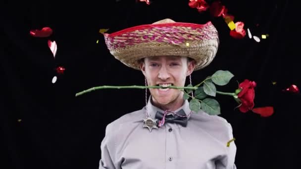 Konfeti, dans ve Rose 'la stüdyoda siyah arka planda bir adam. Parti, kutlama etkinliği ve Meksika sombrero 'sunda erkek müzik eşliğinde dans ediyor, ağzında çiçeklerle eğleniyor ve eğleniyor. - Video, Çekim