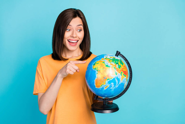 Portrait of positive impressed astonished woman long hairstyle yellow t-shirt look directing at globe isolated on blue color background. - Foto, Imagem