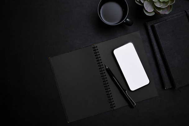 Moderne trendy zwarte werkruimte bovenaanzicht met smartphone wit scherm mockup, pen, notebook, koffiebeker en decor plant op zwarte achtergrond. - Foto, afbeelding