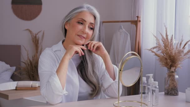 Белая 60-летняя пожилая женщина, 50-летняя женщина с седыми волосами, глядя на зеркальное отражение, избалованная трогательной морщинистой операцией против старения лица, снимает увлажнение кожи лица - Кадры, видео