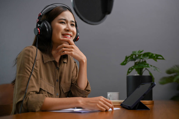 Talented and attractive young Asian female online podcaster or influencer in headphones speaking on her professional microphone, running her online podcast in her home studio. - Photo, Image
