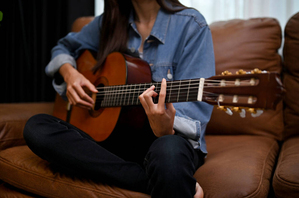 Vonzó fiatal ázsiai nő játszik klasszikus gitáron a nappaliban, az ő vintage barna bőr kanapén. kivágott kép - Fotó, kép