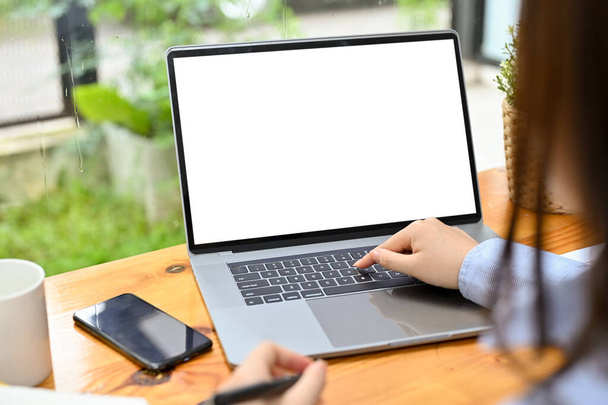 Μια επιχειρηματίας πληκτρολογεί σε ένα πληκτρολόγιο φορητού υπολογιστή, χρησιμοποιώντας ένα φορητό υπολογιστή για να ολοκληρώσει τις εργασίες της. Φορητός υπολογιστής λευκό οθόνη mockup. κοντινή εικόνα - Φωτογραφία, εικόνα