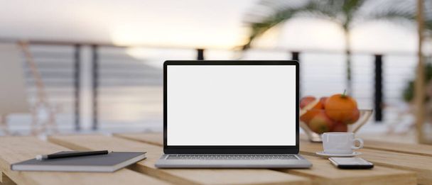 背景のぼやけた海の景色の上に木製のテーブルのノートパソコンの白い画面のモックアップ、コーヒーカップ、スパイラルノートブックやもの。リモートワークと休日のコンセプト3Dレンダリング、 3Dイラスト - 写真・画像