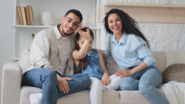 Πολυφυλετική οικογένεια πολυεθνική Ισπανόφωνος Καυκάσιος Αραβες Ινδοί γονείς μητέρα πατέρας με κόρη κορίτσι παιδί κάθεται στον καναπέ στο σπίτι κουνώντας τα χέρια Γεια σας μιλάμε σε απευθείας σύνδεση τηλεδιάσκεψη κλήση - Πλάνα, βίντεο