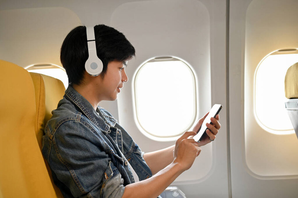 Schöner junger asiatischer männlicher Reisender oder Passagier auf dem Fensterplatz in der Economy Class benutzt während des Fluges sein Handy. - Foto, Bild