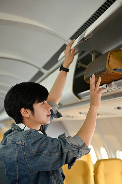 Handsome young Asian male traveler or passenger putting his baggage into an overhead locker. Vacation, traveling, holiday, journey. - Photo, Image