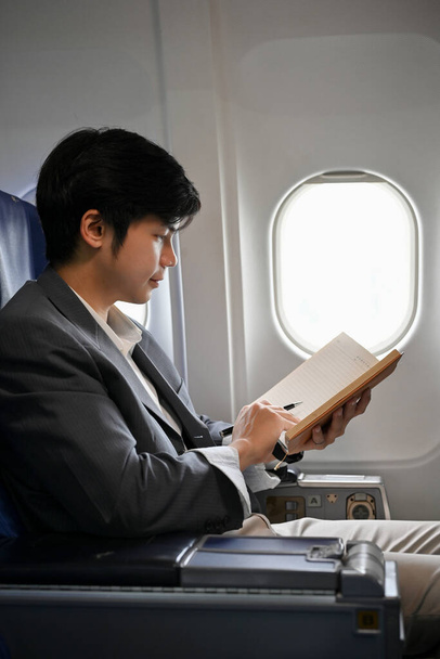 Retrato, Um jovem empresário asiático profissional está no voo, lendo, verificando ou escrevendo algo em seu caderno. - Foto, Imagem