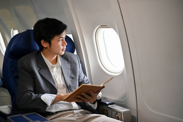 Profesional y exitoso empresario asiático o gerente ejecutivo masculino sosteniendo un libro mientras mira por la ventana, leyendo un libro durante el vuelo para su viaje de negocios. - Foto, Imagen