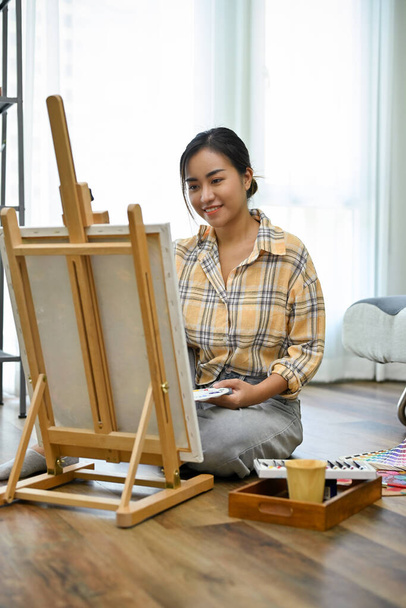 Portrait, Inspired and talented young Asian female artist painting with an acrylic color on her empty canvas easel in her home living room. - Photo, Image