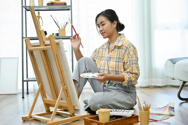 Jolie jeune peintre asiatique assise sur le sol devant la toile et dessinant avec de la couleur acrylique dans son atelier. concept créatif - Photo, image