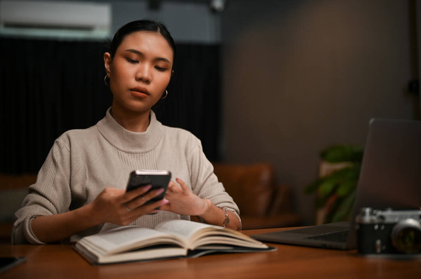Wunderschöne asiatische Millennial-Frau plaudert mit jemandem, während sie nachts entspannt ein Buch in ihrem Wohnzimmer liest. - Foto, Bild