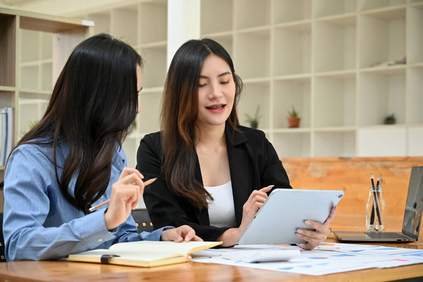 Charming and happy millennial Asian businesswoman showing something on tablet screen, sharing her creative marketing ideas to her female colleague. - Photo, Image
