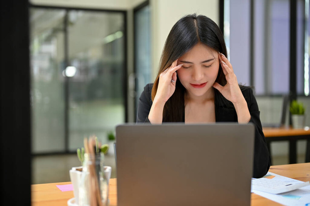 Gestresste und erschöpfte junge asiatische Geschäftsfrau oder Büroangestellte sitzt an ihrem Schreibtisch und spürt Kopfschmerzen oder Migräne.  - Foto, Bild