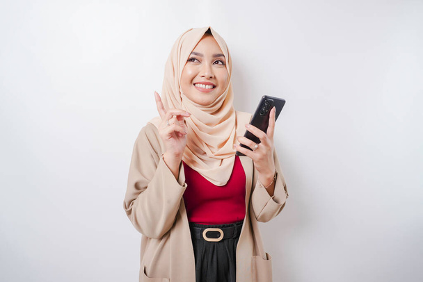 Excited Asian woman wearing hijab pointing at the copy space on top of her while holding her phone, isolated by white background - Φωτογραφία, εικόνα