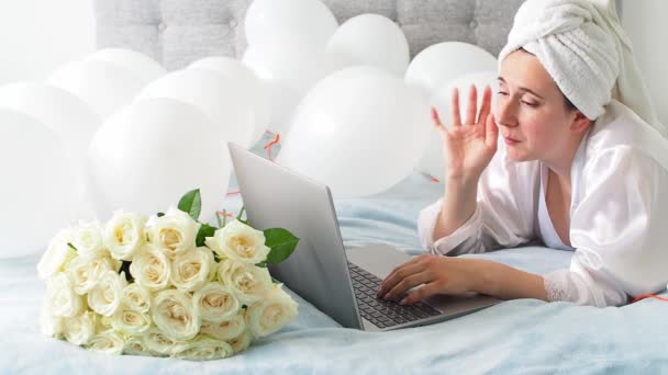 Woman in white clothes and towel on her head lying on a bed with white color balloons and bouquet of roses. Woman works at home, using her laptop, computer. Birthday, wedding. Copy space. Preparation. - Materiał filmowy, wideo