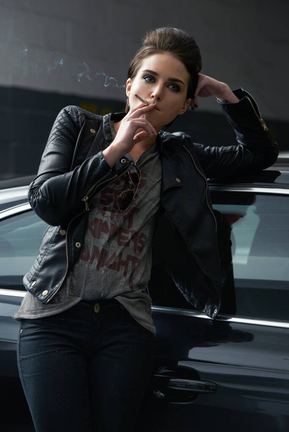 Attitude and attractive. a rebellious looking young woman smoking a cigarette in a car garage - Photo, image