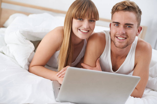 Réservez l'escapade parfaite. Un jeune couple couché au lit travaillant sur un ordinateur portable - Photo, image