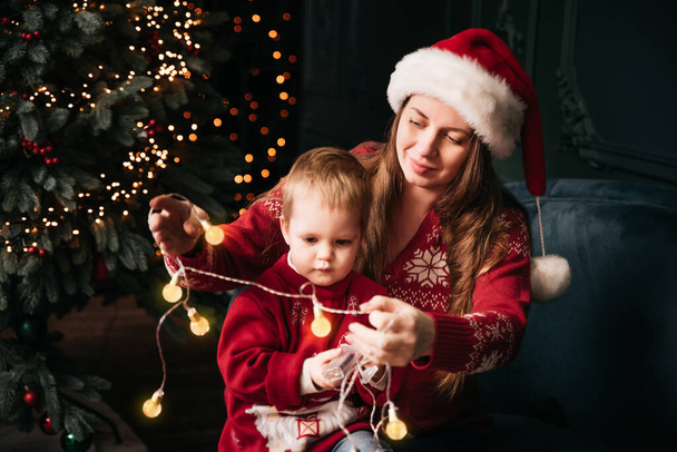 Happy family under Christmas tree. baby boy in Santa Claus hat with gifts under Christmas tree with many gift boxes presents. Happy Holidays, New year. Cozy warm winter evening at home. Xmas time - Foto, Imagen