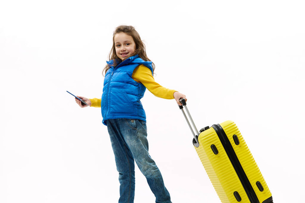 Full length: fashionable little child, traveler girl in blue jeans, yellow shirt and down jacket, traveling abroad for holidays, walking with suitcase on white background. Journey Travel Tourism Trip - Photo, Image