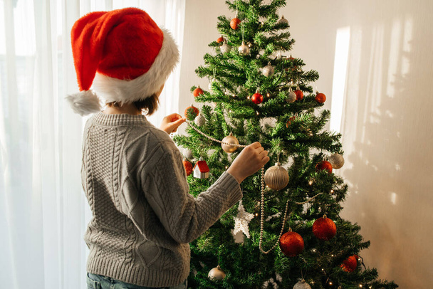 Yılbaşı ve Noel. Noel Baba şapkalı genç bir kız Noel ağacını çelenklerle süslüyor. Tatil için evini dekore ediyorsun.. - Fotoğraf, Görsel